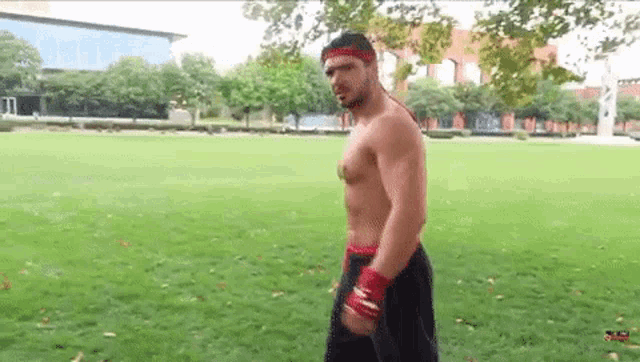 a shirtless man wearing a red headband is standing in a grassy park .