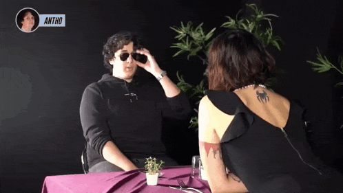 a man and a woman are sitting at a table and the man is wearing sunglasses and the woman is wearing a tattoo on her arm