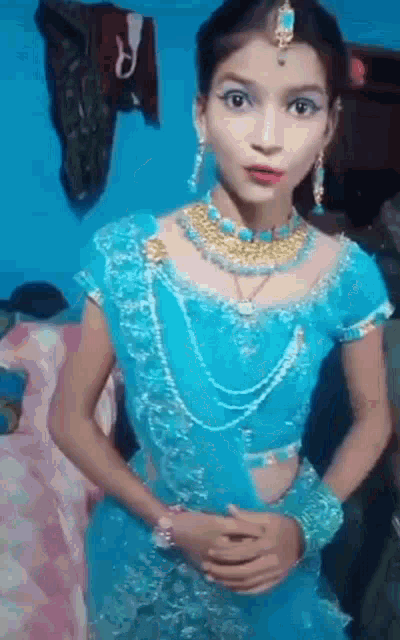 a young girl in a blue dress and jewelry is standing in front of a bed .
