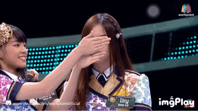 a woman covering another woman 's eyes with her hands and a sign that says bnk 48 on it