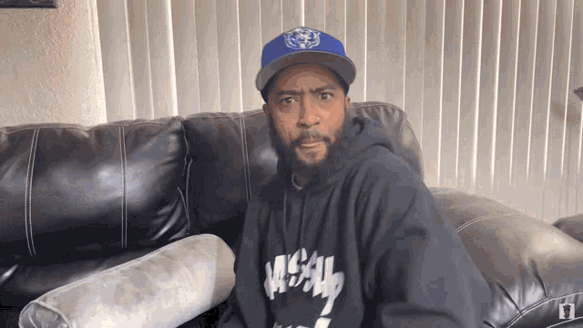 a man sitting on a couch wearing a blue hat and a black hoodie that says mc-al