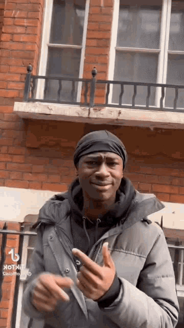 a man in a jacket and hat is standing in front of a brick building and making a funny face .