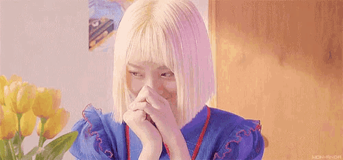 a woman with blonde hair is covering her face with her hands while sitting in front of flowers .