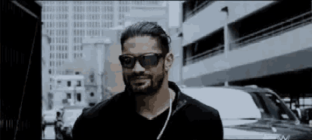 roman reigns is wearing sunglasses and a black shirt in a city .