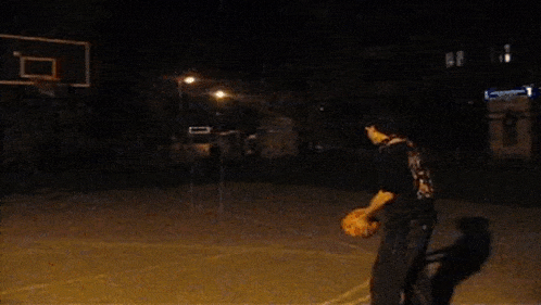 a man is playing basketball at night on a court with a basketball hoop in the background .