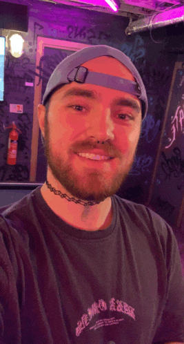 a man wearing a blue hat and a choker smiles in front of a wall with graffiti on it