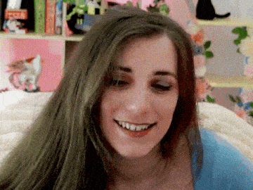 a woman with long green hair is smiling in front of a bookshelf with a cat on it