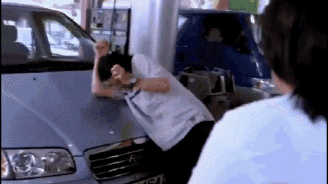 a man is laying on the hood of a silver car while another man looks on .