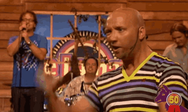 a bald man in a striped shirt stands in front of a group of men singing into microphones