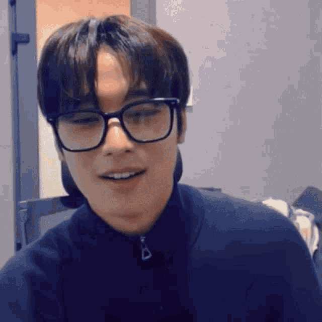 a young man wearing glasses and a black jacket is sitting in a room .