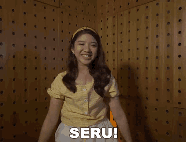a girl in a yellow shirt is standing in front of a wooden wall with the word seru written on it .