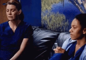 two women in scrubs are sitting on a couch looking at their phones .