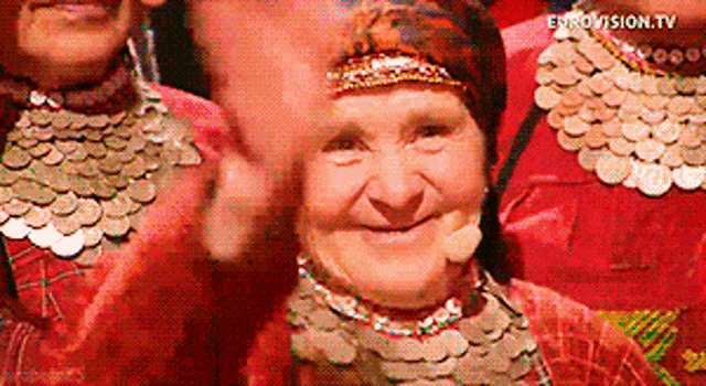 a woman wearing a necklace of coins applauds in front of eurovision tv