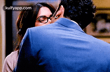 a man and a woman are hugging and kissing . the woman is wearing glasses and the man is wearing a blue suit .