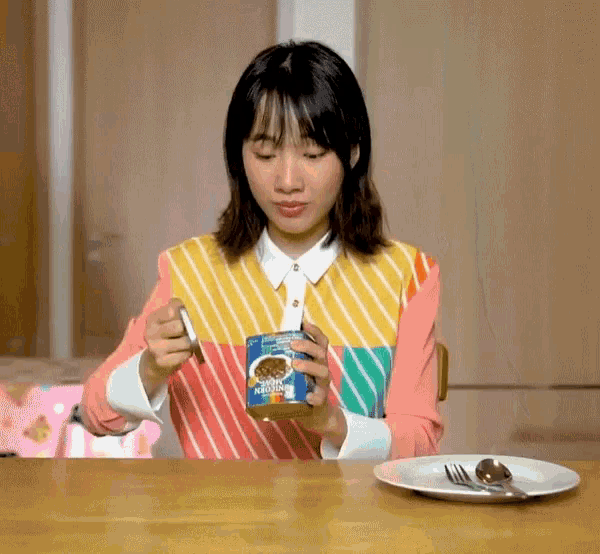 a woman sitting at a table with a plate and spoon holding a can of beans