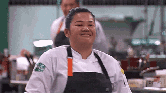 a woman in a chef 's uniform is smiling in a kitchen while a man stands behind her .