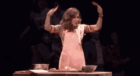 a woman in a pink apron is standing in front of a table with bowls of food and a book .