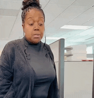 a woman in a black sweater and black shirt is standing in a cubicle in an office .