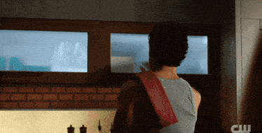 a man in a grey tank top is standing in a kitchen with a cw logo behind him
