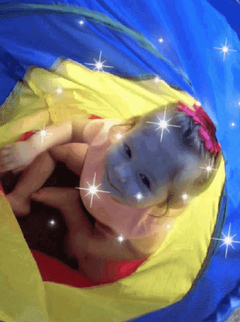 a little girl with a pink flower in her hair is playing in a blue yellow and red tunnel