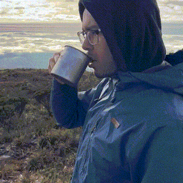 a man wearing glasses and a blue jacket drinks from a mug