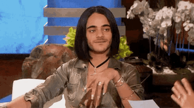 a man with long hair and a beard is sitting at a table with his hands on his hips .