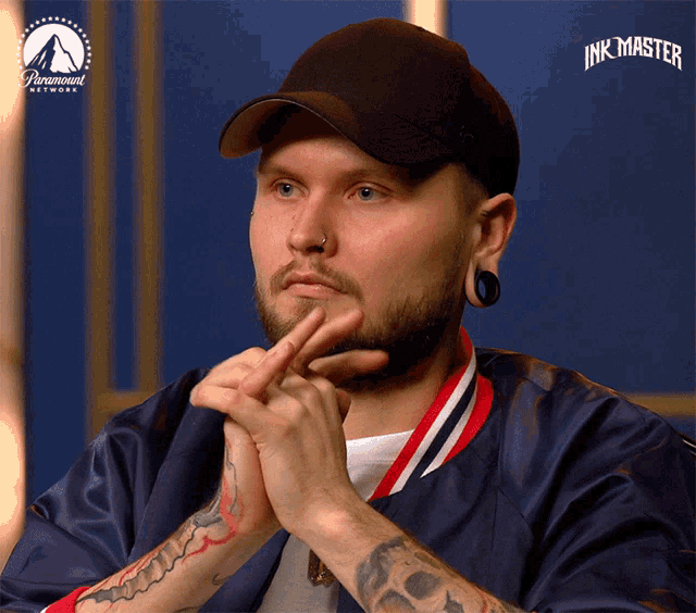 a man wearing a black hat and a blue jacket has his hands folded in front of a screen that says ink master