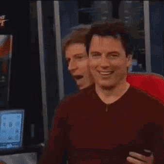 a man in a red sweater is smiling and holding a cell phone while another man looks on .