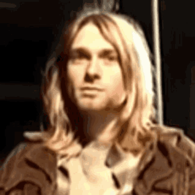 a man with long blonde hair wearing a brown jacket looks at the camera