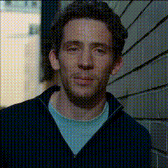 a man standing in front of a brick wall wearing a black sweater and a blue shirt