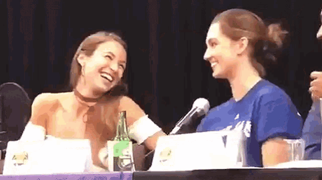 two women are laughing while sitting at a table with a bottle of beer .