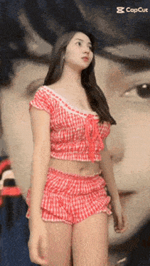 a woman in a red and white plaid top and shorts is standing in front of a painting of a woman 's face