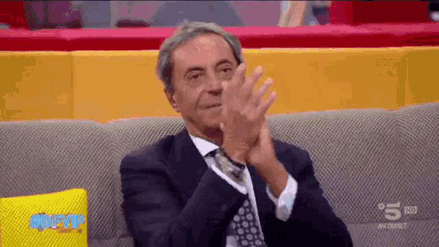 a man in a suit and tie is sitting on a couch clapping his hands