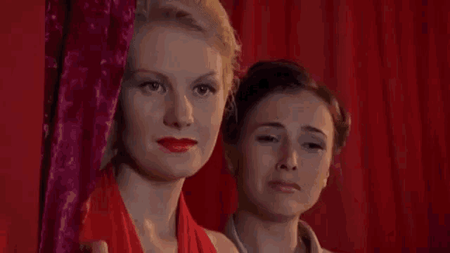 two women standing next to each other in front of a red curtain .