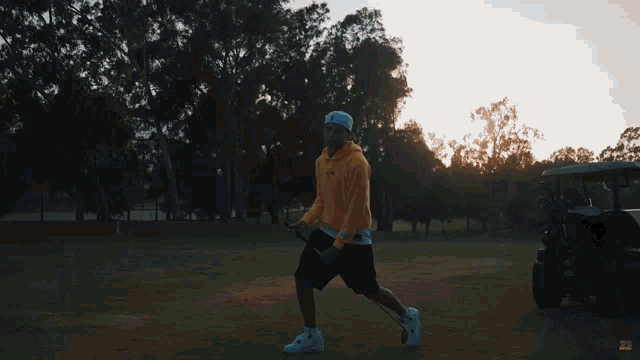a man in a yellow hoodie and white hat is holding a golf club in a park