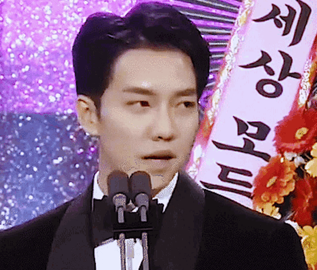 a man in a tuxedo stands in front of two microphones with a sign behind him that says ' a '
