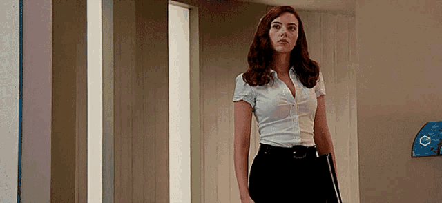 a woman in a white shirt and black pants is standing in a hallway holding a book .