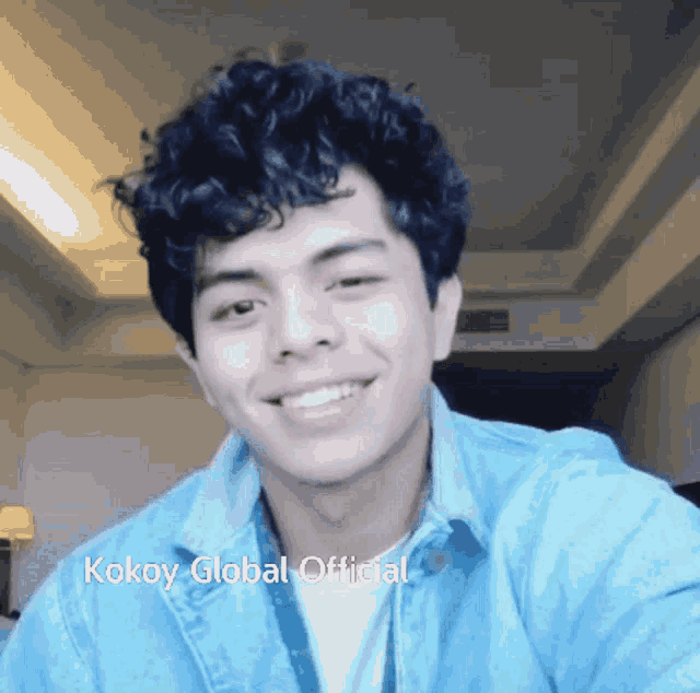 a young man with curly hair is wearing a blue shirt and smiling with the words kokoy global official below him