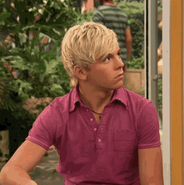 a man with blonde hair wearing a pink shirt looks to the side