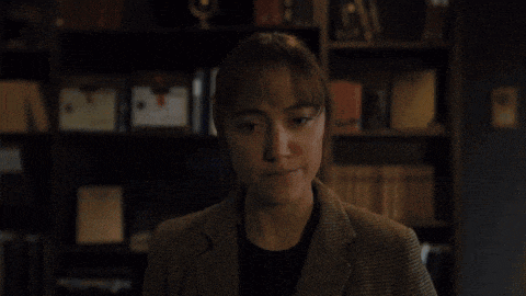 a woman in a suit is sitting in front of a bookshelf in a dark room .