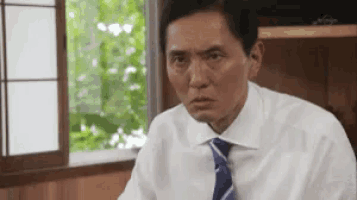 a man in a white shirt and blue tie is sitting in front of a window and making a funny face .