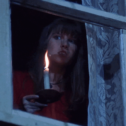 a woman holding a lit candle looks out a window at night