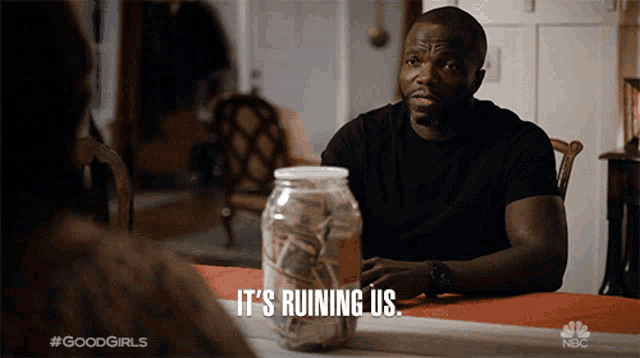 a man is sitting at a table with a jar of money and says it 's ruining us