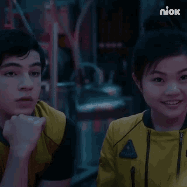 a boy and a girl are sitting next to each other and the girl is wearing a yellow jacket with a triangle on it