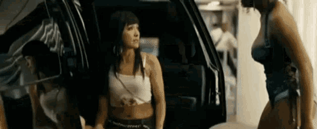 two women are standing next to each other in front of a black car .