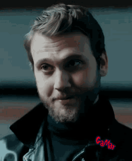 a man with a beard wears a black turtleneck and a leather jacket with gamar written on the sleeve