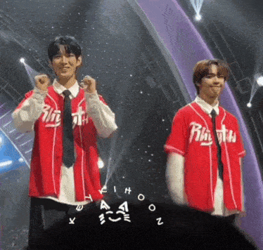 two young men are standing on a stage wearing red baseball jerseys with the word rhythm on them .