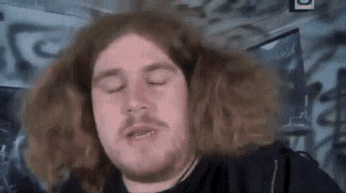 a man with long hair and a beard is looking at the camera with his eyes closed .