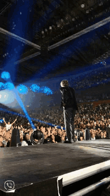 a man in a black jacket stands on a stage in front of a crowd at a concert