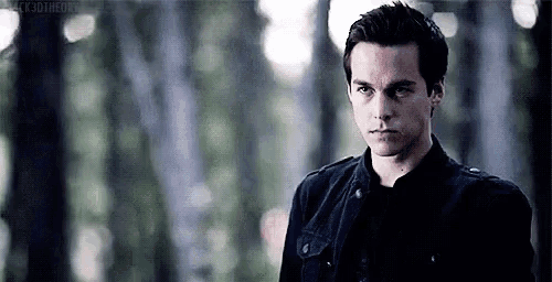 a young man in a black jacket is standing in the woods looking at the camera .